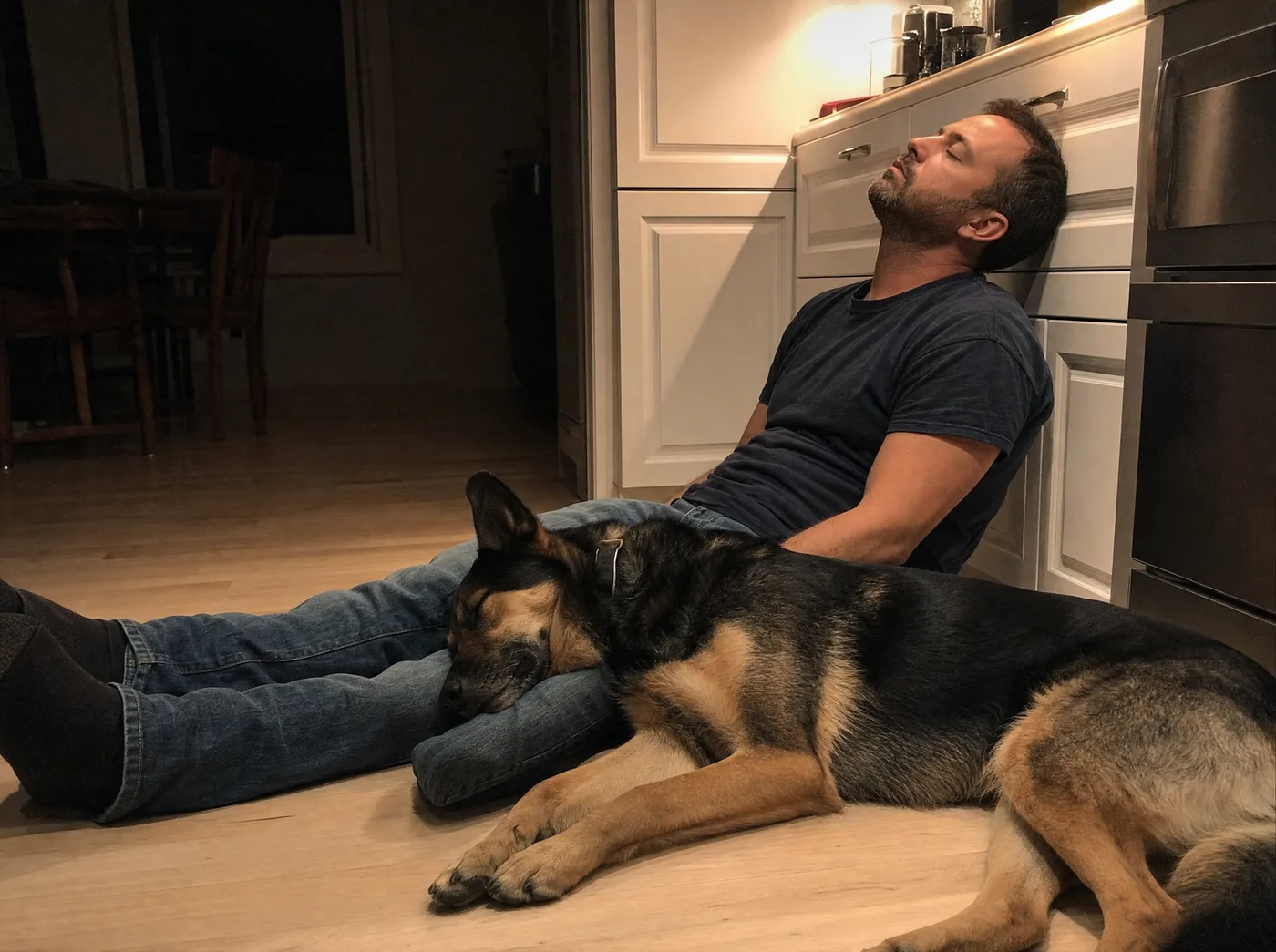 A man asleep against the kitchen cabinets with the dog asleep against his leg